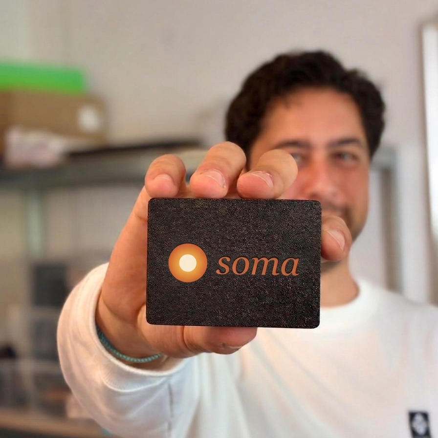 Een hand houdt de Soma-hub vast: een zwart kaartformaat kastje met het Soma-logo in reli&euml;f.